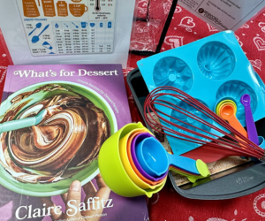 Cooking kit unpacked with recipe book, whisk, muffin molds, pan, and measuring cups.